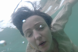 A person's face in close-up, underwater with eyes closed. The water is cold and blue, and water bubbles pass the camera.