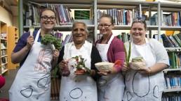 Four women of different ages in aprons holding clay objects including a glazed spoon, a flower-holder and a bowl. They are standing in a library.