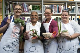 Four women of different ages in aprons holding clay objects including a glazed spoon, a flower-holder and a bowl. They are standing in a library.