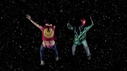 Two figures in colourful clothing leap into a dark sky filled with stars