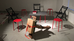 An image of around 8 chairs painted black and red, sited in a gallery space around an strange cardboard construction