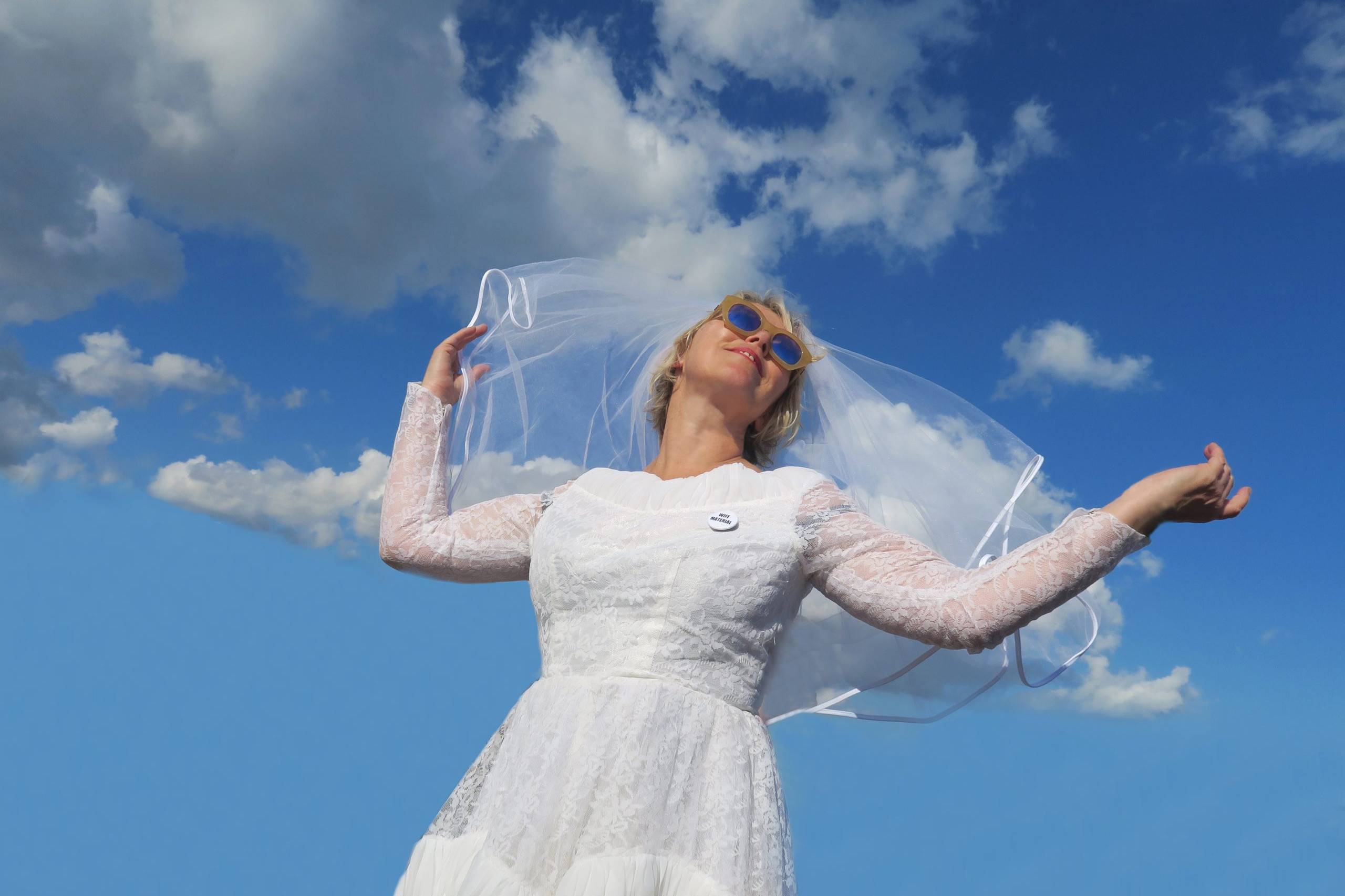 An image of a white-veiled woman in a bridal dress, wearing sunglasses and looking to the horizon against a blue sky