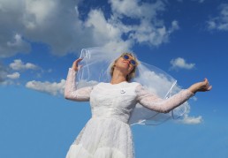 An image of a white-veiled woman in a bridal dress, wearing sunglasses and looking to the horizon against a blue sky
