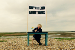 A blonde woman sits behind a desk, which is located on a shingle beach. Above her head is a large sign, reading: Boyfriend Auditions