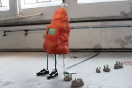 Bright orange paper mache blob like sculpture with stick legs wearing football boots