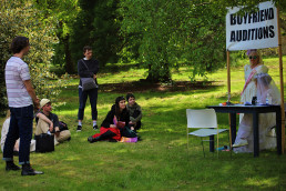 Group pf people sitting on grass looking at two women sitting under a sign with which reads Boyfriend Auditons
