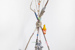 a tripod sculpture made with sticks, feathers and bits hang off the sticks on string