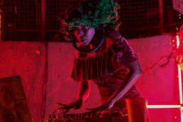 A woman wearing a red crushed velvet dress, large green textured wig and headphones hung around her neck is DJing in a room filled with red light.⁠